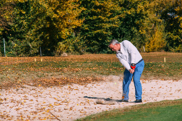 A senior golfer hitting his ball out of a bunker with the sand and autumn background