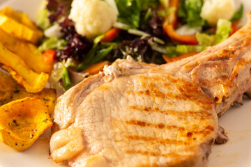 Fried pork entrecote on the bone with stewed pumpkin pieces and salad in a plate close up, green salad with cauliflower and napa cabbage, bell pepper and spices