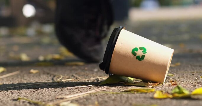 A man walks past a disposable paper cup with a "recycle" sign lying on the ground. People`s desregard towards the garbage that can be recycled.