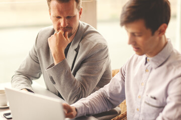 Two businessmen sitting at table in cafe and discussing business presentation on laptop screen, selective focus on middle aged man with his hand on chin