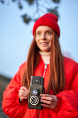 redhead woman in red coat holding retro camera in hands, focus on camera. photography concept