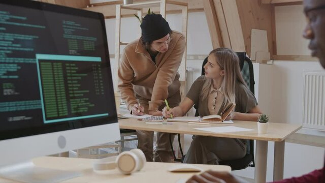 Panning of young Caucasian woman and man discussing new design project while young African male program developer sitting at workplace and coding on computer