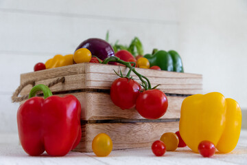 Une caisse de légumes fraiche avec des tomates, des oignons,des poivrons,des carottes,des tomates cerises avec de belles couleurs éclatantes rouges, jaunes,vert et oranges sur un fond de bardage blanc