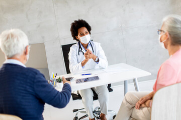 Obraz premium Senior couple with protective facial masks receive news from black female doctor