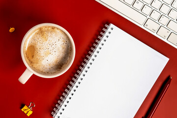 Top view of the office desk. A workspace with an empty notebook, keyboard, stationery, and coffee cup on a red background.