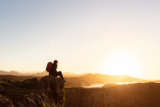 Side view of anonymous male hiker with backpack sitting on rock in mountains and enjoying amazing sundown during vacation