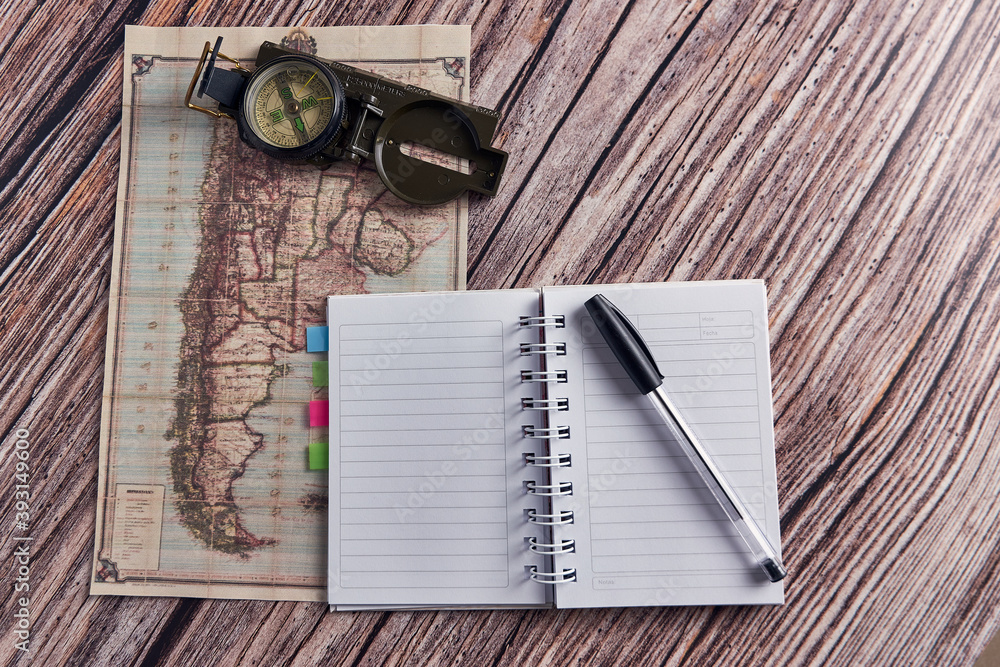 Poster top view closeup of a compass placed on an ancient map and a notebook on a wooden table