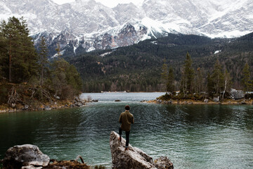Back view of man standing on stone on shore of pond and observing mountains covered with snow in winter