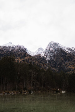 Dark lake and forested mountains with snow cowered rocky peaks under gray cloudy sky in cold autumn day