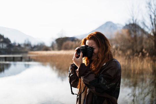 Young female photographer in warm jacket standing on coast of lake against blurred mountains and taking pictures with photo camera in autumn day