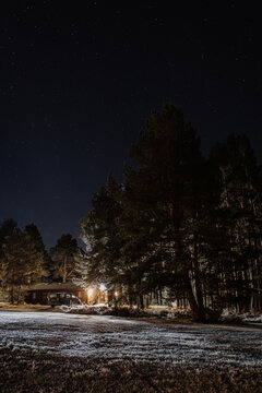 Amazing scenery of dark snowy forest with illuminated houses at night under starry sky in Scottish Highlands