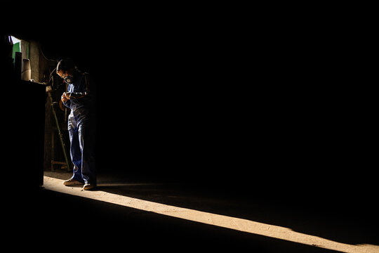 Side view of unrecognizable mechanic in protective respirator and uniform standing in dark garage and illuminated by sunbeam