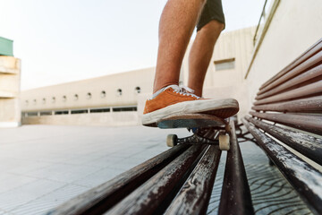 Low angle of unrecognizable crop male standing on skateboard on wooden bench and showing stunt