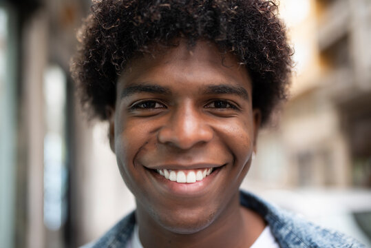Positive African American male with Afro hairstyle and charismatic smile looking at camera while standing on blurred background of city