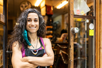 Positive adult ethnic female mechanic in colorful apron with protective mask smiling and looking at camera with arms crossed standing on entrance of bike service workshop