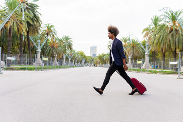 Side view of a young black man in a suit dragging a wheeled suitcase down the street on a tree-lined avenue