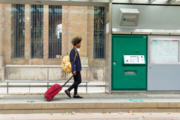 Side view of black man with afro hair walking on a tram station with a suitcase on wheels and a bag hanging from his shoulder