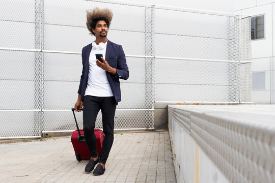 Black man in a suit with a wireless headphones around his neck and a suitcase walking while using the mobile outdoors