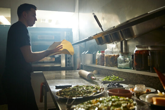 Side View Of Man Flipping And Tossing Dough On Table With Flour While Stretching Pizza Base In Restaurant Kitchen