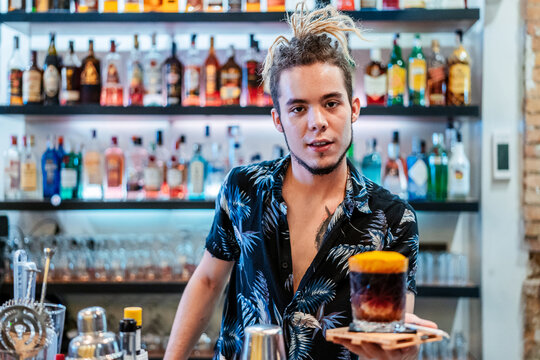 Stylish male bartender with dreadlocks standing at counter in bar with cold alcohol cocktail and looking at camera