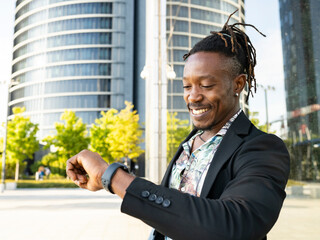 Low angle side view of positive African American male entrepreneur looking at wristwatch and checking time while standing in city and waiting for meeting