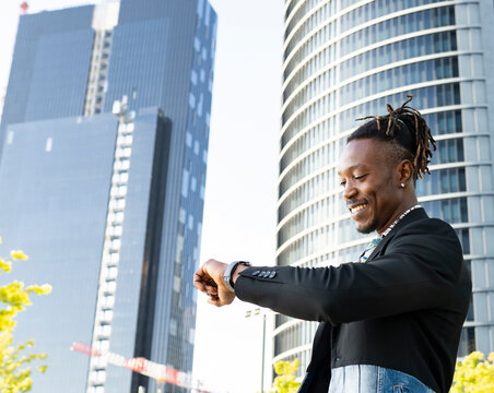 Low angle side view of positive African American male entrepreneur looking at wristwatch and checking time while standing in city and waiting for meeting