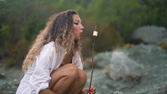 Model with prefect tanned skin holds stick with marshmallow and blows on hot dessert squatting against large grey stones and bushy green trees closeup slow motion.