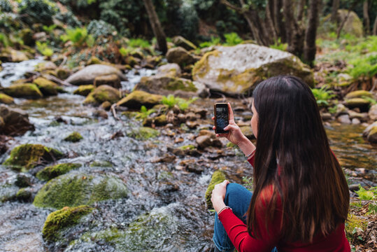 Back view of anonymous female tourist sitting on shore of river in woods and taking picture of amazing landscape in Valle del Jerte
