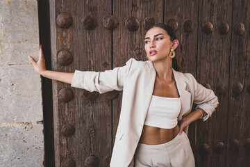Delightful slim ethnic female model with perfect makeup wearing stylish light suit standing against weathered wooden door of urban building
