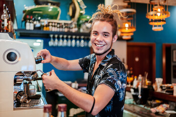 Side view of cheerful male barista with dreadlocks preparing aromatic coffee in coffee maker while looking at camera