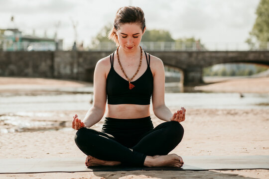 Tranquil female in sportswear sitting on mat in Padmasana with mudra hands and meditating while practicing yoga with closed eyes on beach