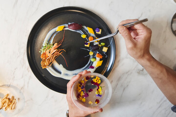 From above of crop anonymous chef with tongs arranging colorful edible blossoms on plate while garnishing sophisticated dish in restaurant kitchen