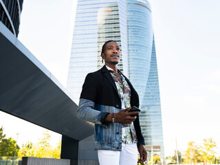 Low angle of stylish African American male executive manager standing with cellphone on street in city center and looking away
