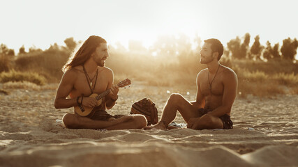 Relaxed talented man playing acoustic guitar for male friend while sitting together on sandy beach at sunset