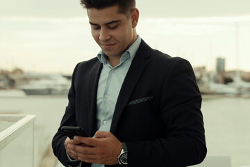 Smiling male entrepreneur in formal suit standing on the street of city and texting on smartphone