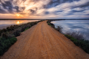 dirt road that divides the waters, on the left the sunset with orange colors and on the right clouds and blue tones