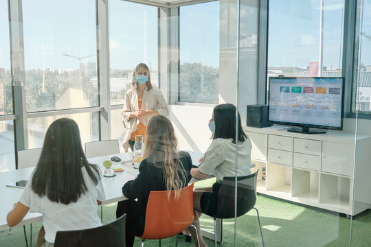 Young female colleagues in protective masks with notebooks in hands standing near window and discussing business issues while gathering together in contemporary workspace during business meeting