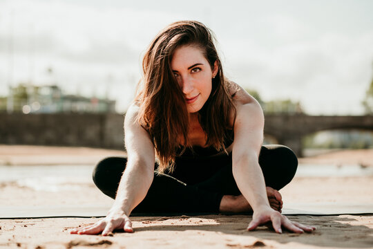 Smiling flexible female sitting on mat in Janu Sirsasana and doing forward bend while looking at camera and practicing yoga on beach in summer