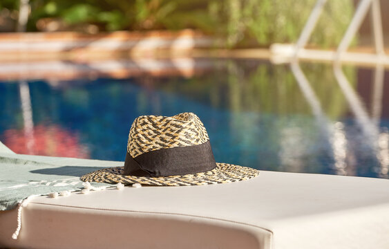 Elegant straw hat with brown band placed on lounge chair at poolside in tropical hotel in summer day