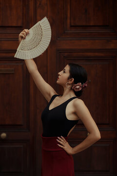 Side view of Hispanic female Flamenco dancer with white fan performing passionate dance near wooden door