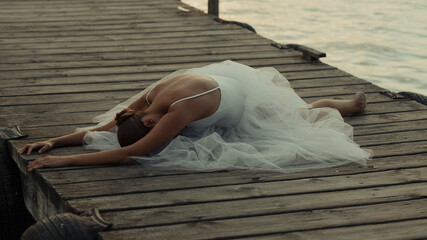 Unrecognizable elegant female ballet dancer in white tutu performing graceful seated pose on wooden pier near sea at sunset time