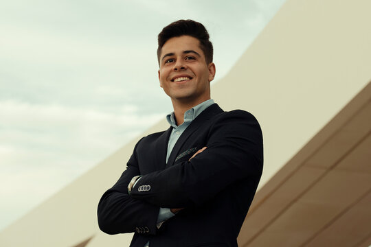 Content handsome businessman standing with crossed arms near building in city and looking away
