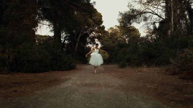 Full body talented young female ballet performer in white dress and pointe shoes dancing on path in green forest in summer evening