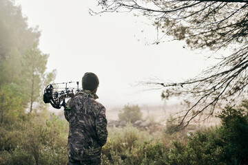 Back view of unrecognizable man in camouflage standing with compound bow in forest and looking away during hunting