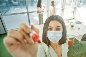 Young female member of business team in protective mask taking notes on glass wall while working with colleagues and searching for solution of business problem in contemporary office