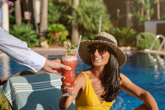Cheerful young female tourist in stylish straw hat and sunglasses receiving fresh cold fruit beverage from unrecognizable barmen while enjoying summer vacation at poolside in resort