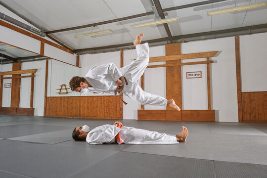 Side view of two people with mask and kimono practicing krav maga while one jumps on top of the other in a gym