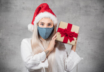 Fototapeta na wymiar Surprised beautiful blond woman wears christmas santa claus hat - cap and holding red gift box - present with ribbon, isolated on gray background. A Corona - Covid 19 Christmas concept