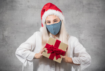 Santa woman shows christmas gift isolated on gray background. Happy young girl wearing red santa hat and holding present box. A Corona - Covid 19 Christmas concept