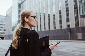 Rear view of businesswoman with in-ear headphones holding file in city